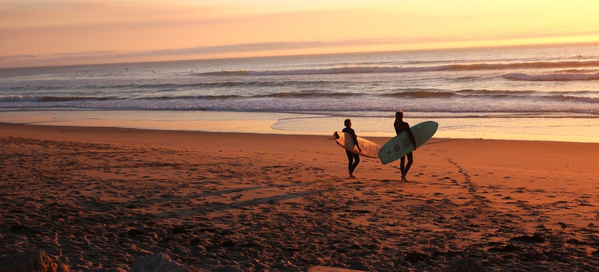 Two surfers with surfboards walking toward the ocean at sunset.